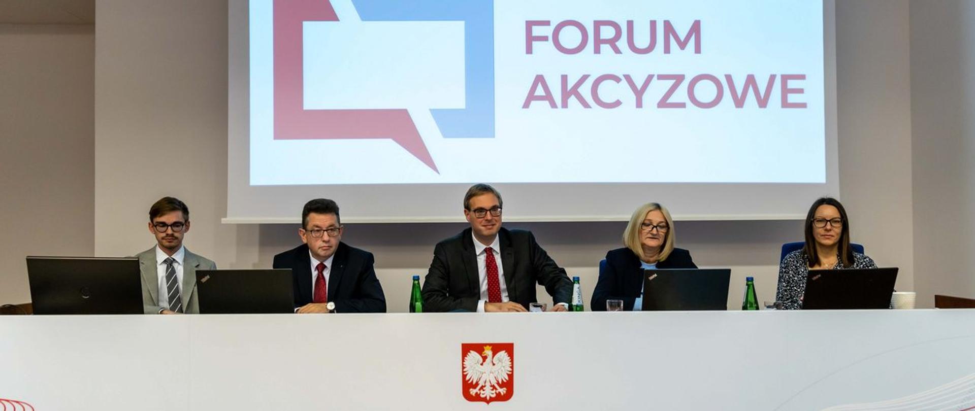 Stół prezydialny, uczestnicy spotkania, w tle wyświetlony napis Forum Akcyzowe.