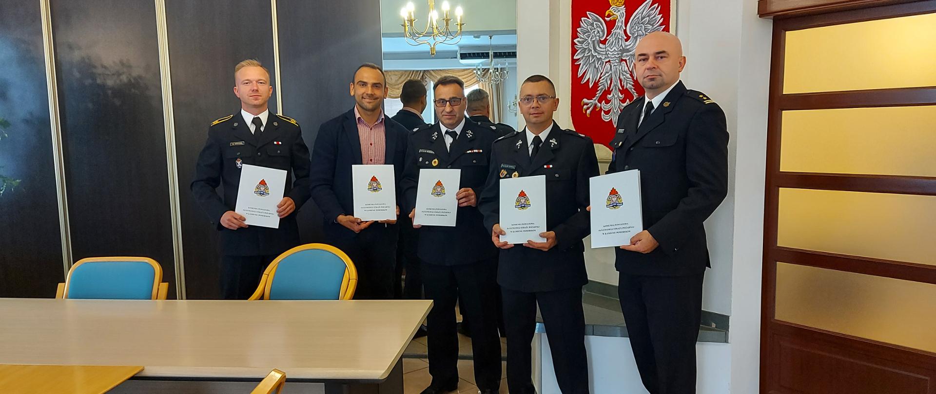 Przedstawiciele straży pożarnej o raz miasta stoją z podpisanymi dokumentami w pomieszczeniu biurowym.