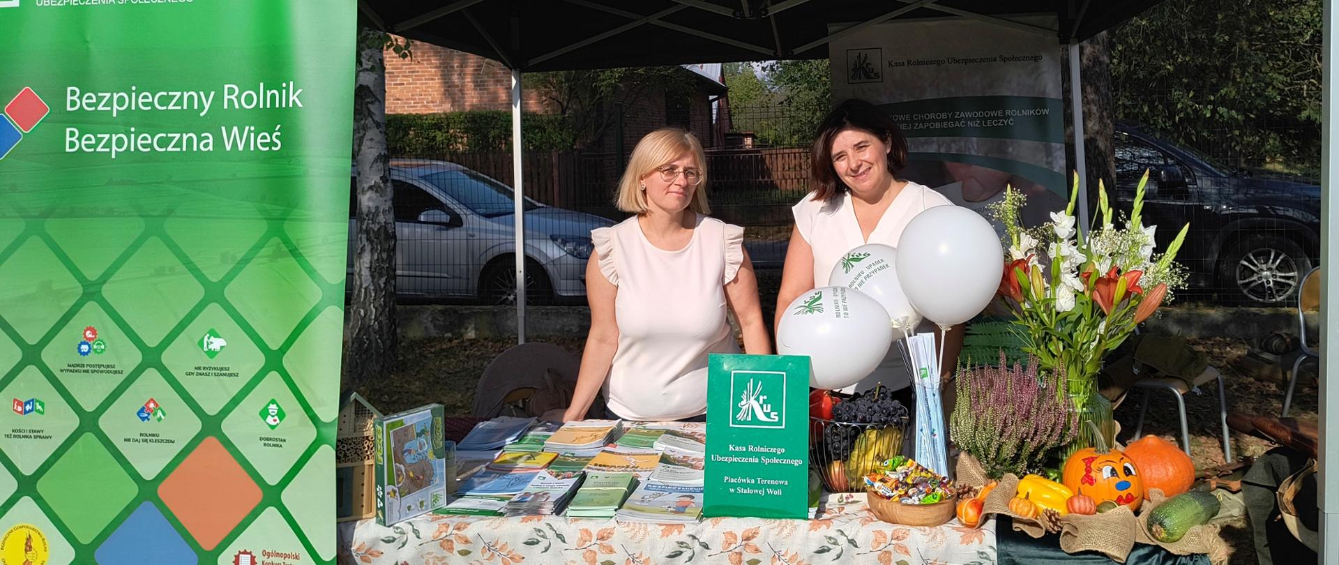 Przy stoisku informacyjno-promocyjnym Kasy znajdują się dwie osoby – pracownice KRUS. Przed nimi znajduje się stół na którym położone są materiały prewencyjne Kasy (ulotki, odblaski) oraz kwiaty i dynie.