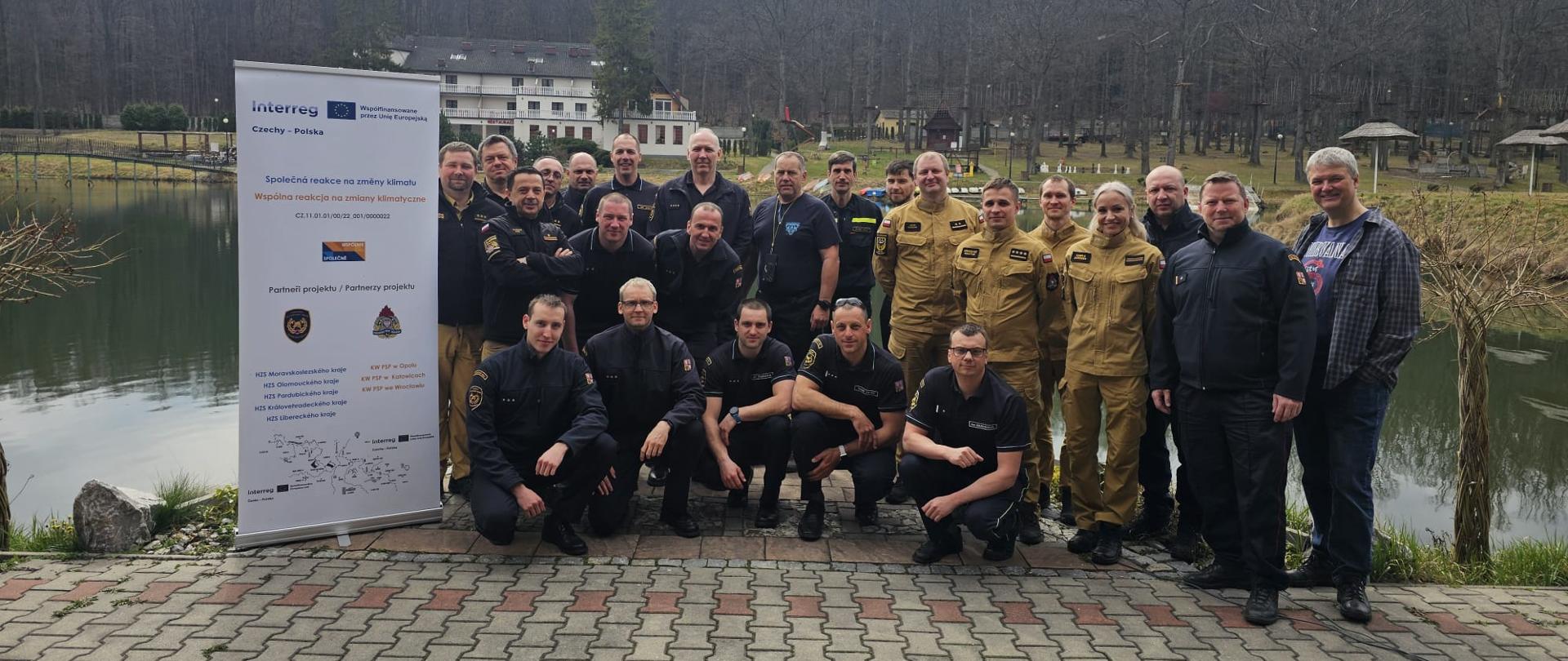 Uczestnicy warsztatów z Polski i Czech pozują do wspólnego zdjęcia przy zbiorniku wodnym podczas wizyty terenowej pierwszego dnia warsztatów dotyczących reagowania na skutki zmian klimatycznych. W tle widoczne są zabudowania i zalesiony teren. Obok grupy stoi baner projektu Wspólna reakcja na zmiany klimatyczne.