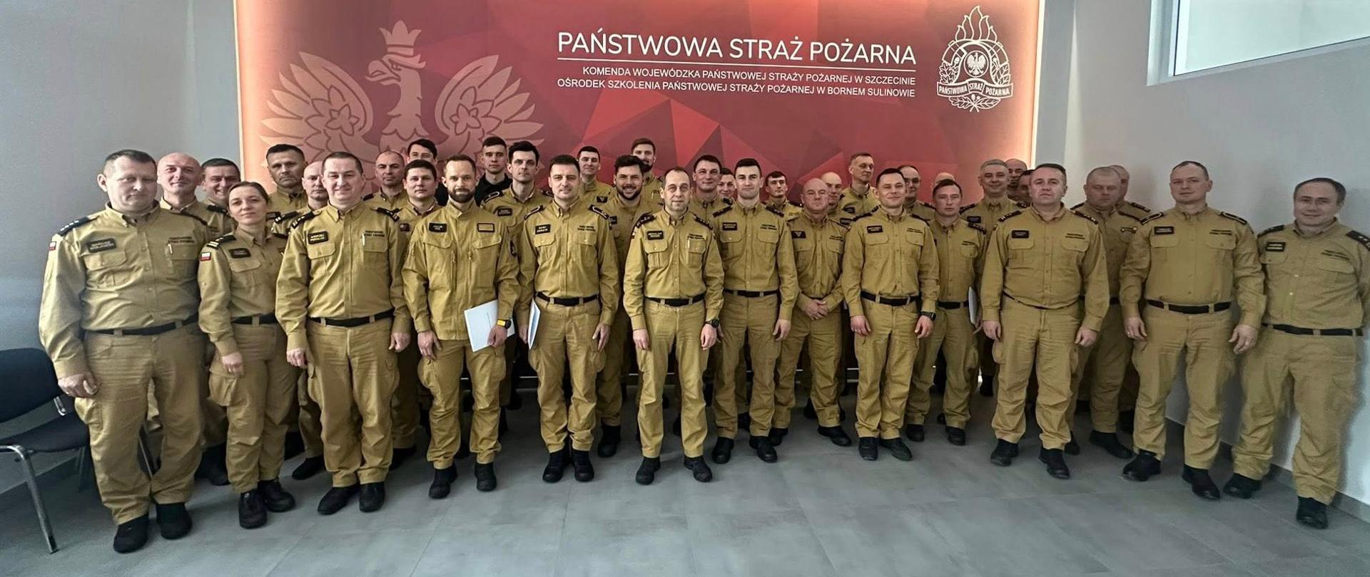 Na zdjęciu widać dużą grupę około trzydziestu strażaków w jednakowych, jasnobrązowych mundurach służbowych. Stoją w kilku rzędach w jasnym, nowoczesnym pomieszczeniu. Wszyscy patrzą w stronę aparatu.
Za nimi znajduje się duża, podświetlana tablica w kolorze czerwonym z napisem „Państwowa Straż Pożarna” oraz godłem i symbolem straży pożarnej. Pomieszczenie ma jasne ściany, sufit z kwadratowymi panelami oraz szarą podłogę. Zdjęcie ma charakter oficjalny i grupowy.