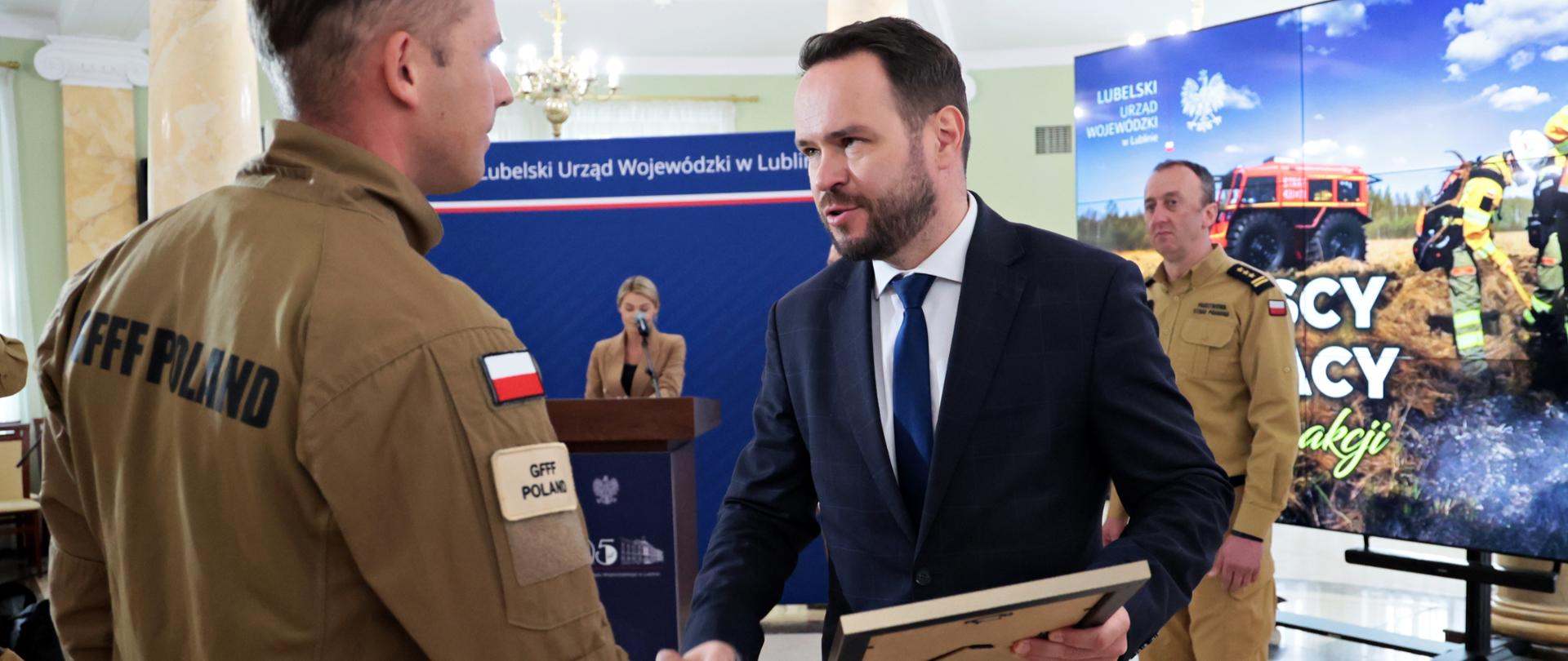 Dwóch mężczyzn w różnym wieku i strojach ściska sobie dłonie na pierwszym planie. Mężczyzna po lewej ma na sobie beżowy mundur z naszywką z polską flagą na ramieniu i napisem "GFFF POLAND". Mężczyzna po prawej jest ubrany w ciemny garnitur, białą koszulę i niebieski krawat (to wojewoda lubelski). Trzyma w ręku płaski, prostokątny przedmiot, prawdopodobnie dyplom lub nagrodę. W tle widoczni są dwaj inni mężczyźni, kobieta za mównicą z herbem Polski oraz duży ekran wyświetlający sceny z akcji ratowniczej i logo "LUBELSKI URZĄD WOJEWÓDZKI". W tle widać również fragment pomieszczenia z oknem i zasłonami.