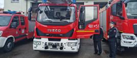 Samochody gaśnicze stoją przed budynkiem drzwi samochodu drabiny mechanicznej są otwarte w środku pojazdu siedzi strażak obok drzwi stoi dwóch funkcjonariuszy Państwowej Straży Pożarnej.