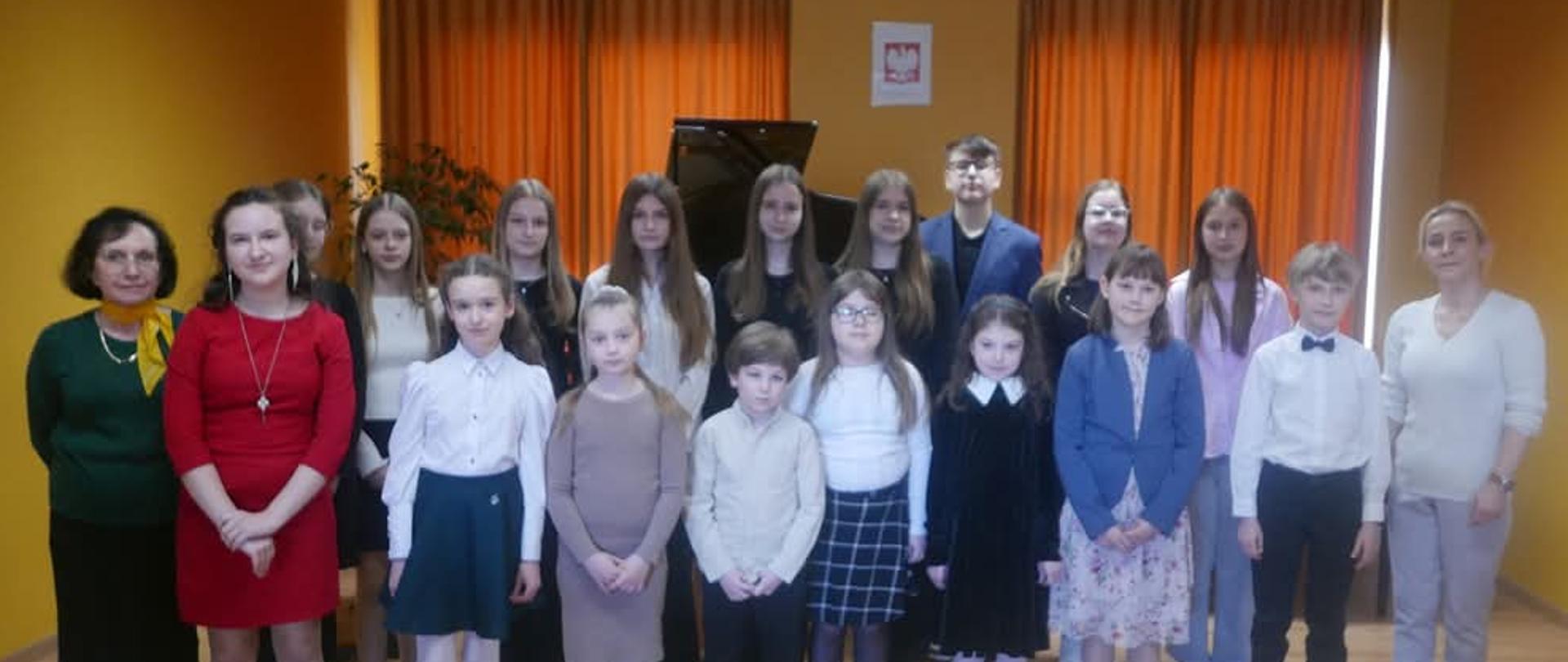 Pamiątkowa fotografia wszystkich grających na koncercie fortepianowym razem z pedagogami. Za nimi dwa fortepiany i zasłonięte kotarami okna.