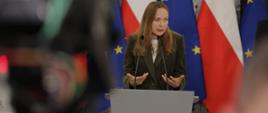 Minister funduszy i polityki regionalnej Katarzyna Pełczyńska-Nałęcz stoi za mównicą z mikrofonami, za jej plecami flagi Polski i UE