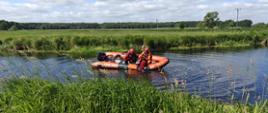 Zdjęcie przedstawia scenę ćwiczeń prowadzonych przez strażaków, na niewielkiej rzece lub kanale. W centralnej części obrazu znajduje się pomarańczowo-szary ponton ratunkowy. W pontonie widać trzech ratowników ubranych w kombinezony i hełmy. Dwóch z nich siedzi, a jeden, ubrany na pomarańczowo, jest częściowo zanurzony w wodzie przy burcie pontonu, co sugeruje, że albo właśnie wchodzi do wody, albo jest w trakcie wykonywania jakiegoś zadania w wodzie. Na burcie pontonu widoczny jest napis "ZODIAC" oraz "PSP", co potwierdza, że jest to sprzęt Państwowej Straży Pożarnej. W pontonie widać również elementy wyposażenia, być może plecak ratowniczy. Rzeka ma ciemną wodę z widocznymi falami i refleksami światła. Brzegi rzeki są gęsto porośnięte wysoką, zieloną trawą i inną roślinnością, tworząc naturalne, wiejskie otoczenie. W tle, za polami, rozciąga się linia lasu. Niebo jest częściowo zachmurzone, z białymi i szarymi chmurami, ale ogólnie dzień jest jasny i słoneczny.