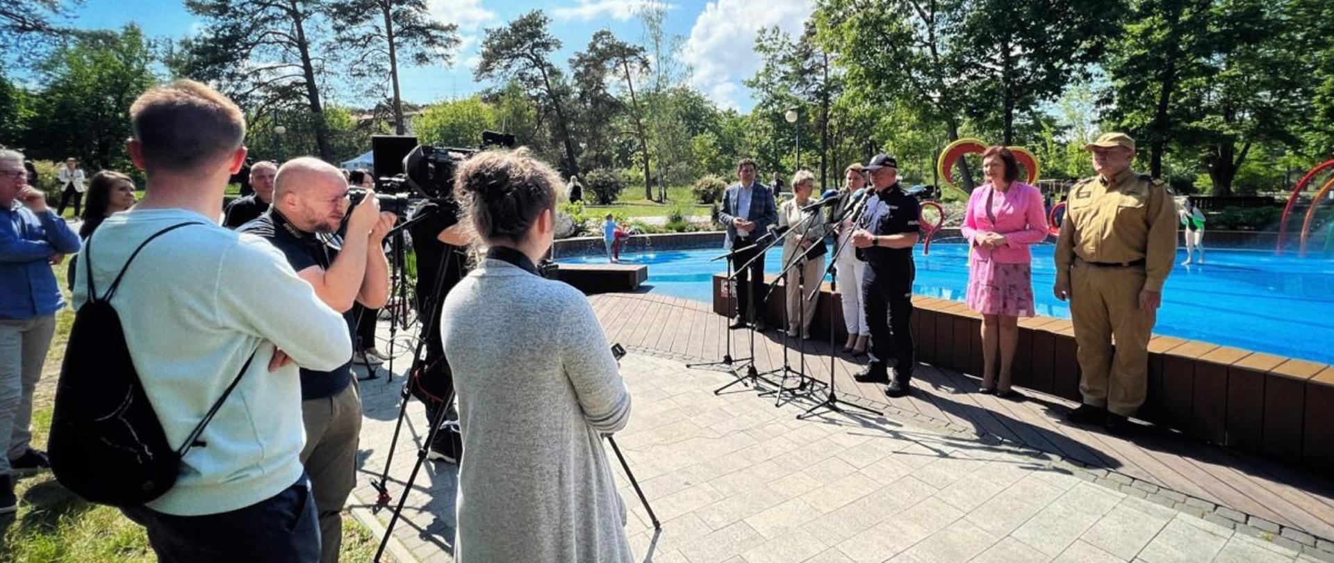 Zdjęcie wykonane na zewnątrz. Konferencja prasowa. Przed mikrofonami stoi sześć osób. Wśród nich funkcjonariusz Państwowej Straży Pożarnej w stopniu generała.