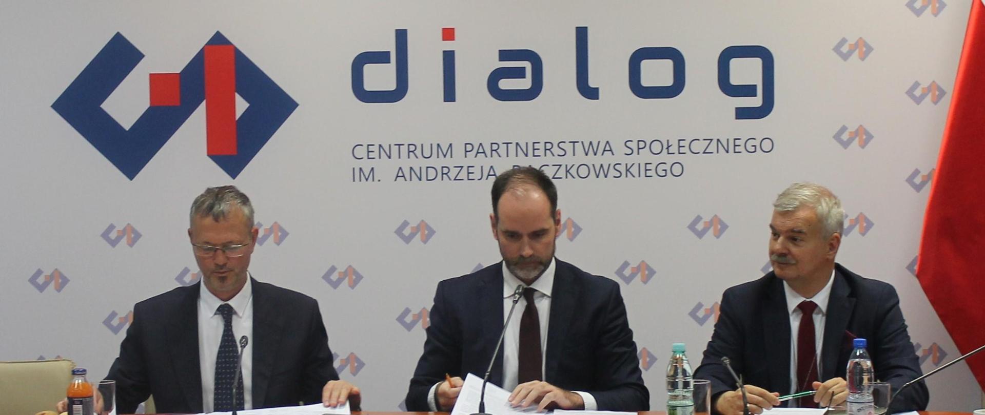 Na zdjęciu widnieje stół prezydialny za którym siedzą: Pan Jacek Bartmiński, Podsekretarz Stanu w Ministerstwie Aktywów Państwowych; Pan Ignacy Niemczycki, Podsekretarz Stanu w Ministerstwie Rozwoju i Technologii oraz Pan Krzysztof Zaręba, Zastępca Dyrektora Departamentu Innowacji i Polityki Przemysłowej w Ministerstwie Rozwoju i Technologii. W tle znajduje się baner z napisem Centrum Partnerstwa Społecznego „Dialog” in. Andrzeja Bączkowskiego.