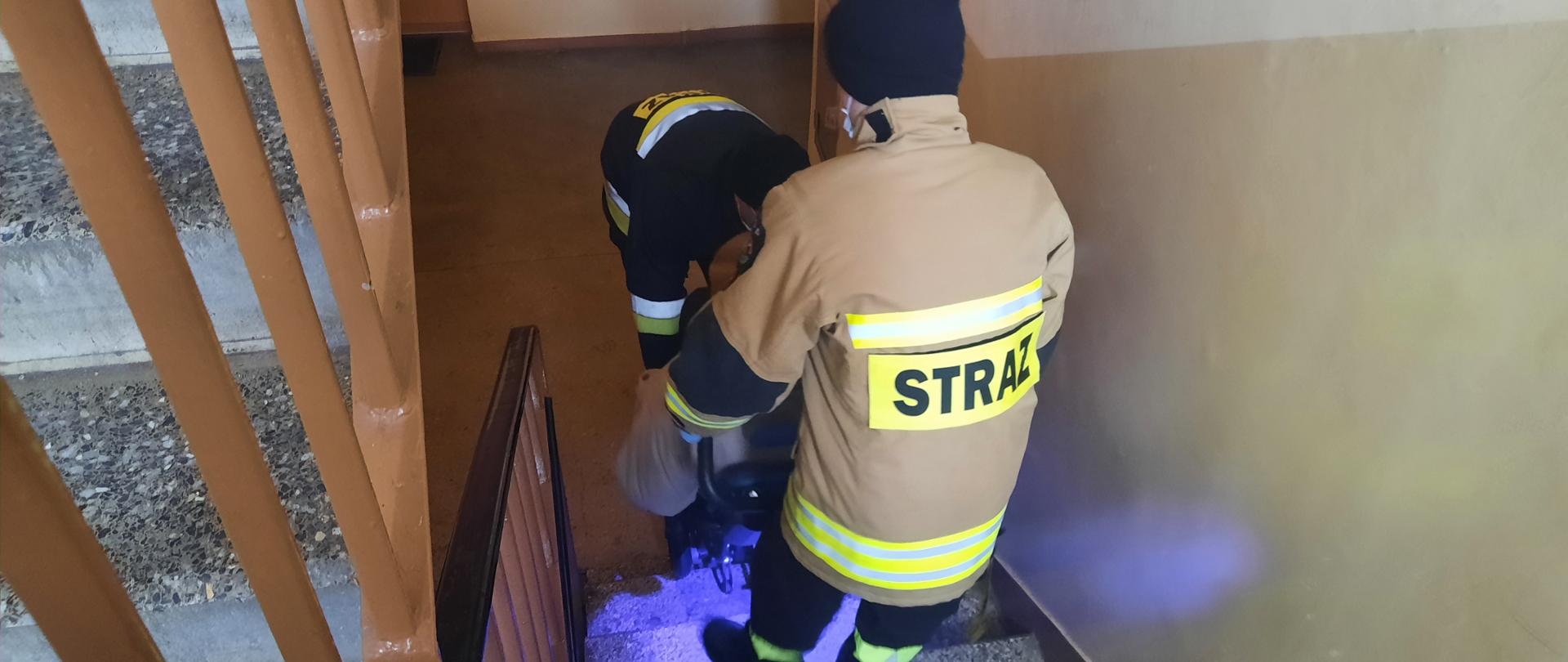 Strażacy pomagaja w transporcie osobie starszej na wózku na szczepienie. 