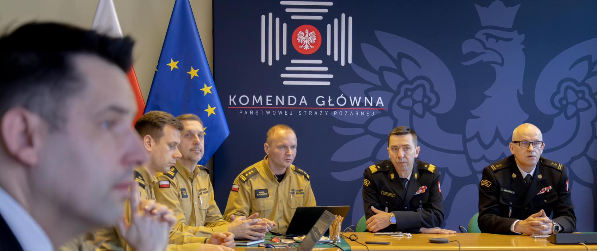 Wnętrze sali konferencyjnej przy stole siedzą strażacy, którzy rozmawiają ze sobą.