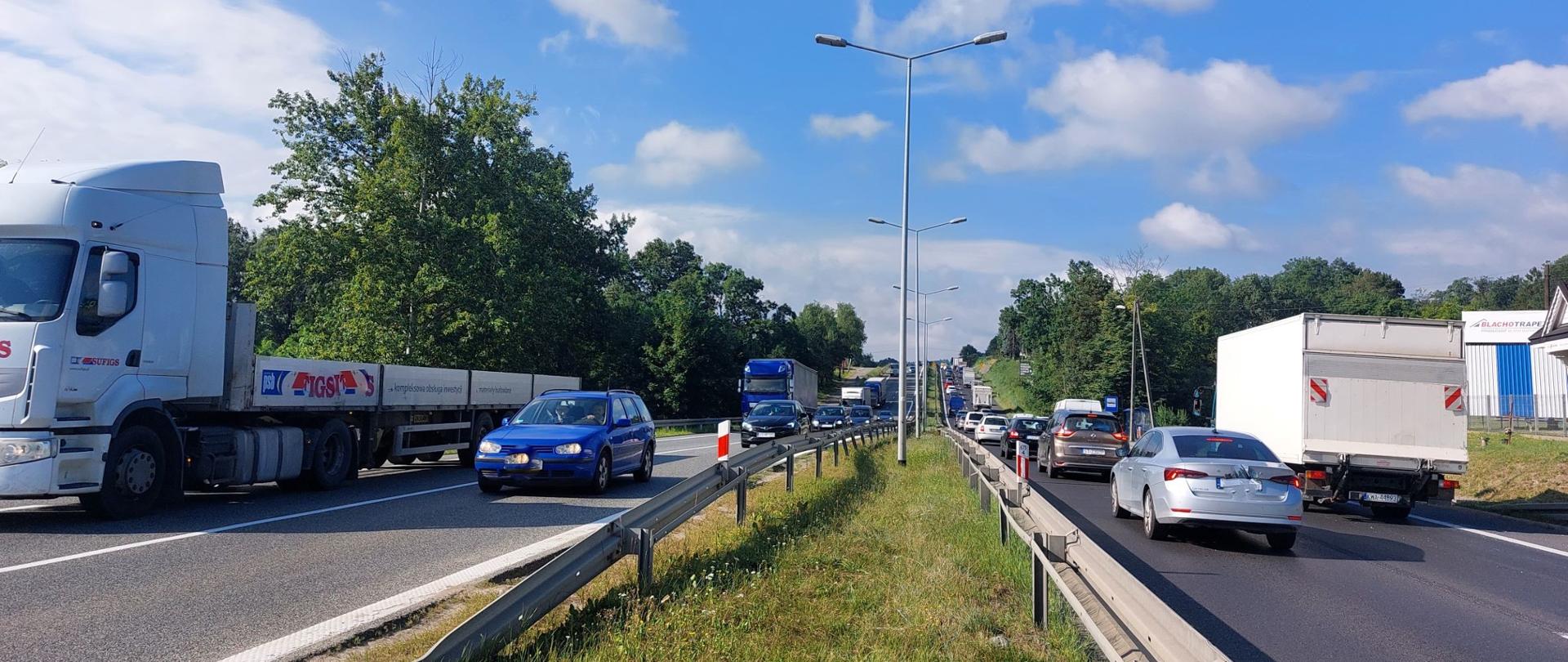 Zdjęcie zrobione ze środka pasa zieleni, który oddziela dwie jezdnie po dwa pasy ruchu w każdym kierunku. Widoczny duży ruch.