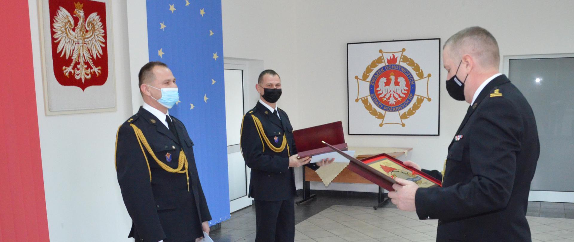 Trzech strażaków stojących na środku, po lewej na ścianie godło RP i flagi unii europejskiej oraz Polski. W tle na ścianie symbol Związku Ochotniczych Straży Pożarnych RP.