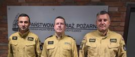 Trzech funkcjonariuszy Państwowej Straży Pożarnej pozuje do pamiątkowego zdjęcia w siedzibie Komendy Miejskiej PSP w Siemianowicach Śląskich. Od lewej: mł. kpt. Michał Janus, Komendant Miejski PSP bryg. Marcin Gąsowski oraz kpt. Sebastian Karpiński. Dwaj mianowani funkcjonariusze trzymają czerwone teczki z decyzjami o mianowaniu.