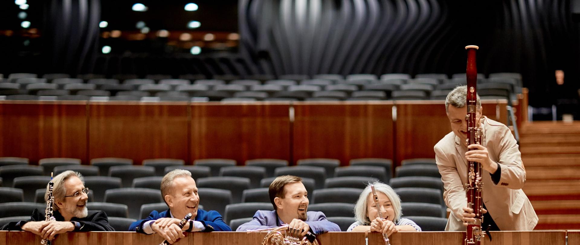 Kolorowe zdjęcie sali koncertowej - część z krzesłami dla publiczności, na środkowych miejscach zasiadają uśmiechnięci członkowie Krakowskiego Kwintetu Dętego, oparci o drewnianą balustradę i prezentujący na niej instrumenty, na których grają.