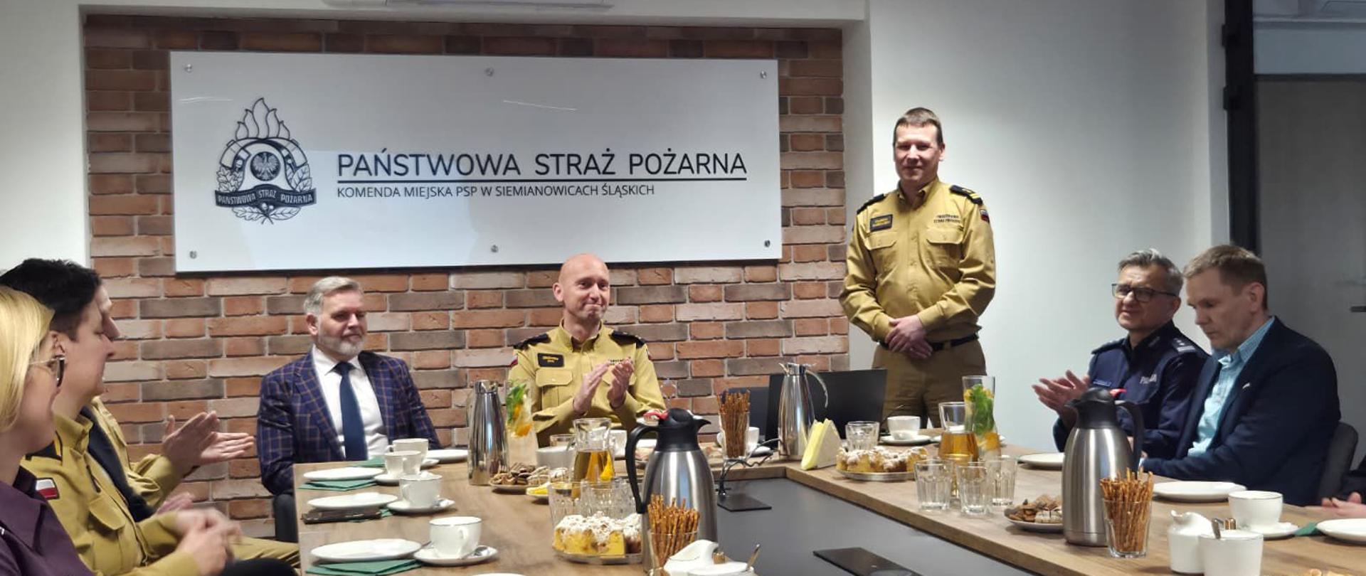 Podczas spotkania w sali konferencyjnej jeden ze strażaków stoi i przemawia do zgromadzonych. Pozostali uczestnicy, siedzący przy stole konferencyjnym, klaszczą. Na stole znajdują się filiżanki, dzbanki z napojami oraz poczęstunek. W tle widoczna jest tablica z nazwą Komendy Miejskiej PSP w Siemianowicach Śląskich.