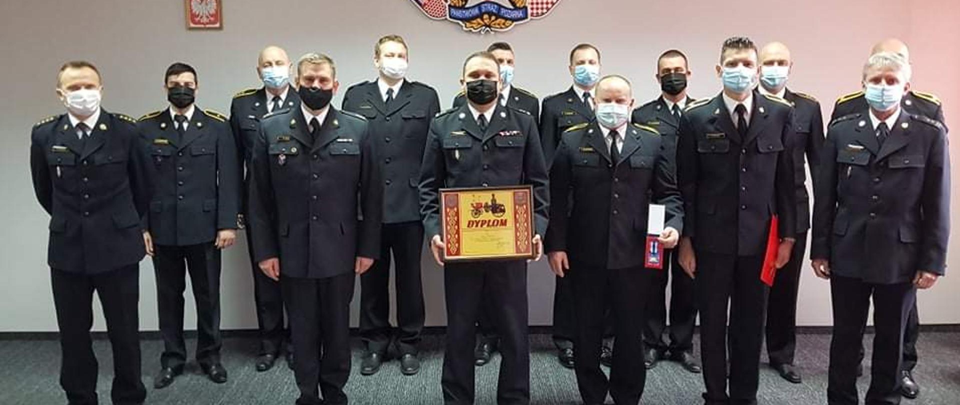 Na zdjęciu widać grupę wyróżnionych i awansowanych funkcjonariuszy KM PSP w Kaliszu. Na środku, z dyplomem stoi Komendant Miejski Państwowej Straży Pożarnej w Kaliszu bryg. Sławomir Kotoński. Za osobami widać ścianę pomieszczenia w kolorze szarym, na ścianie godło państwowe i logo PSP oraz miasta i powiatu kaliskiego. Osoby stoją na ciemnym dywanie. Na suficie kasetony, białe.
