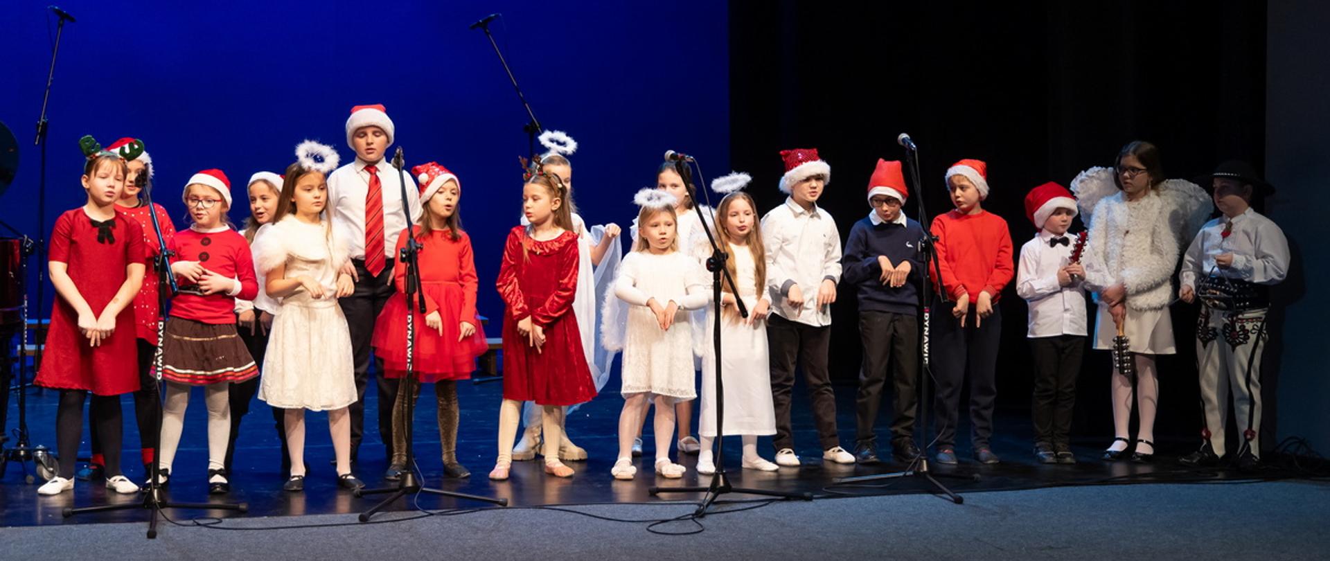 Zdjęcie grupowe, na scenie uczniowie ubrani w świąteczne ubrania stoją w dwóch rzędach. Przed nimi statywy z mikrofonami.