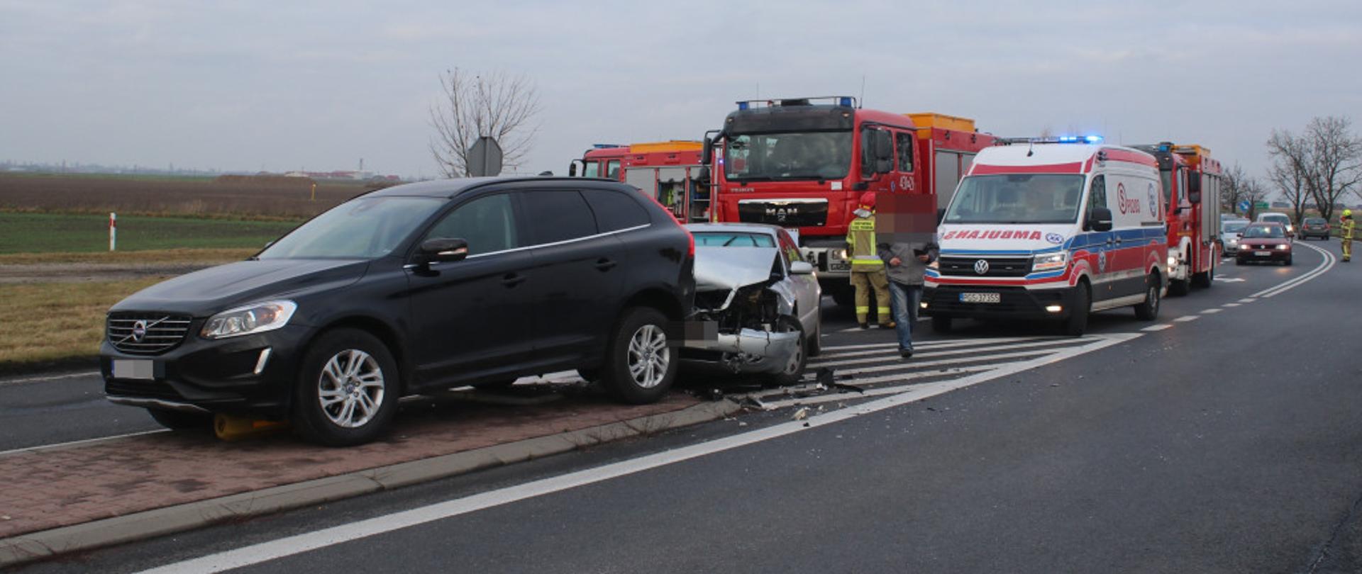 Zdjęcie przedstawia samochody osobowe, które uległy uszkodzeniu. W tle stoją służby ratownicze.