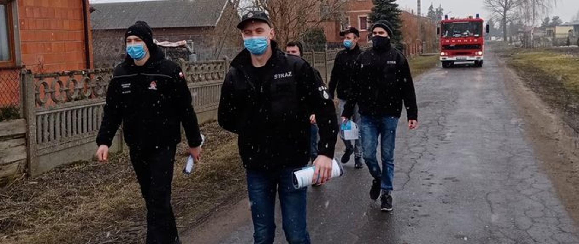 Na zdjęciu widać druhów z OSP w Sławęcinie, którzy idą drogą do gospodarstwa zostawić ulotkę. W tle widać zabudowania oraz samochód strażacki.