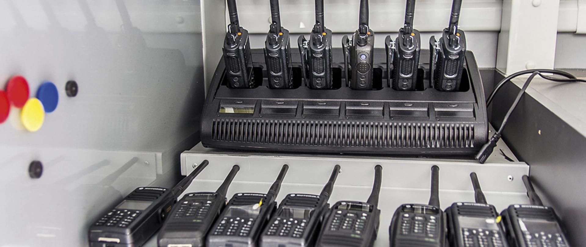 Sześć radiostacji nasobnych w stacji ładującej. Na pierwszym planie osiem sztuk radiostacji leżących na stoliku