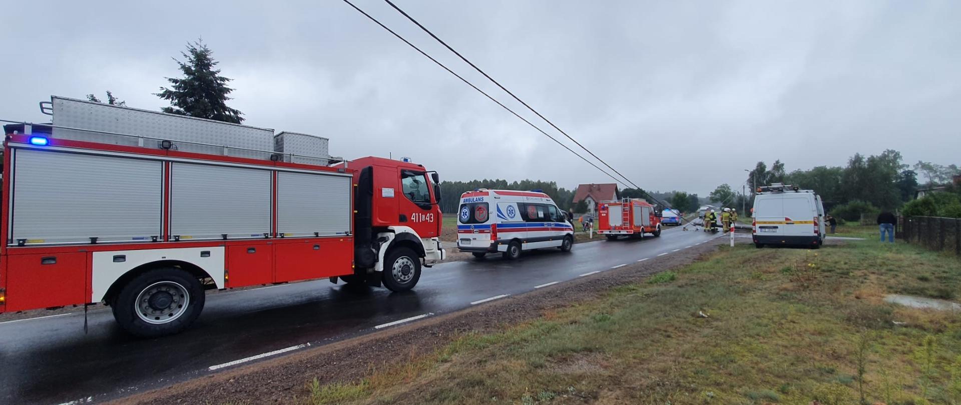 Niebo zachmurzone całkowicie, na pierwszym plamie widoczny od prawy bok czerwonego samochodu Straży Pożarnej zaparkowany na drodze asfaltowej z włączoną niebieską sygnalizacją ostrzegawczą, na prawych drzwiach numer operacyjny 411-43. Przed samochodem Straży Pożarnej stoi biała karetka pogotowia, przed karetką pogotowia stoi drugi czerwony samochód Straży Pożarnej. W oddali widać biały samochód dostawczy Pogotowia Energetycznego oraz grupę strażaków ubranych w ubrania specjalne. Przez całość zdjęcia widać nisko zawieszone przewody energetyczne. W dalekim kadrze zdjęcia widać srebrny radiowóz Policji oraz Przewrócony na drogę słup energetyczny. 