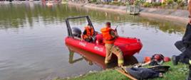 Na zdjęciu widoczna jest łódź ratownicza Państwowej Straży Pożarnej. Obok niej znajduje się funkcjonariusz Państwowej Straży Pożarnej. W łódce znajduje się jeden funkcjonariusz Państwowej Straży Pożarnej. Łódź jest zwodowana na zalewie ośrodka wypoczynkowego "GUTWIN".