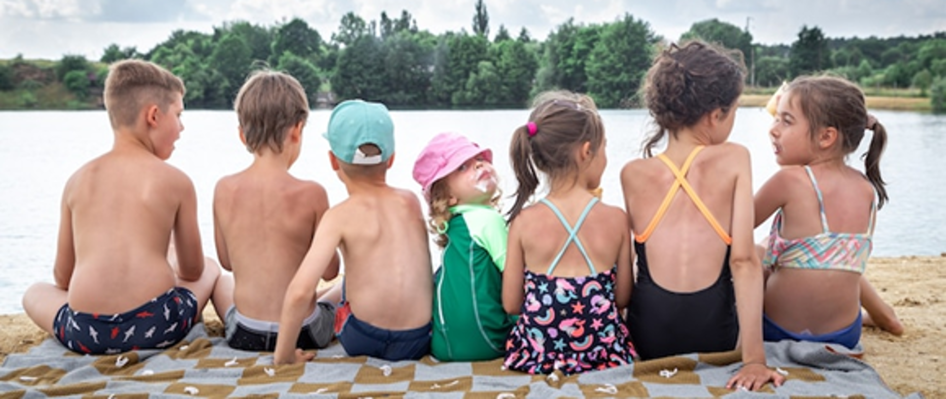 Widok z tyłu dzieci. Siedzą nad rzeką i odpoczywają po kąpieli, opalają się, jedzą lody.