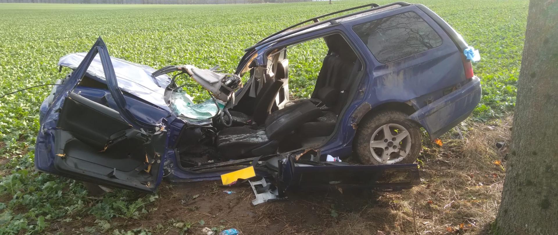 Zdjęcie przedstawia rozbity niebieski samochód osobowy, który ma wyciętą część karoserii. Auto znajduje się na polu rzepaku. Samochód ma zmiażdżoną przednią maskę od uderzenia w drzewo .