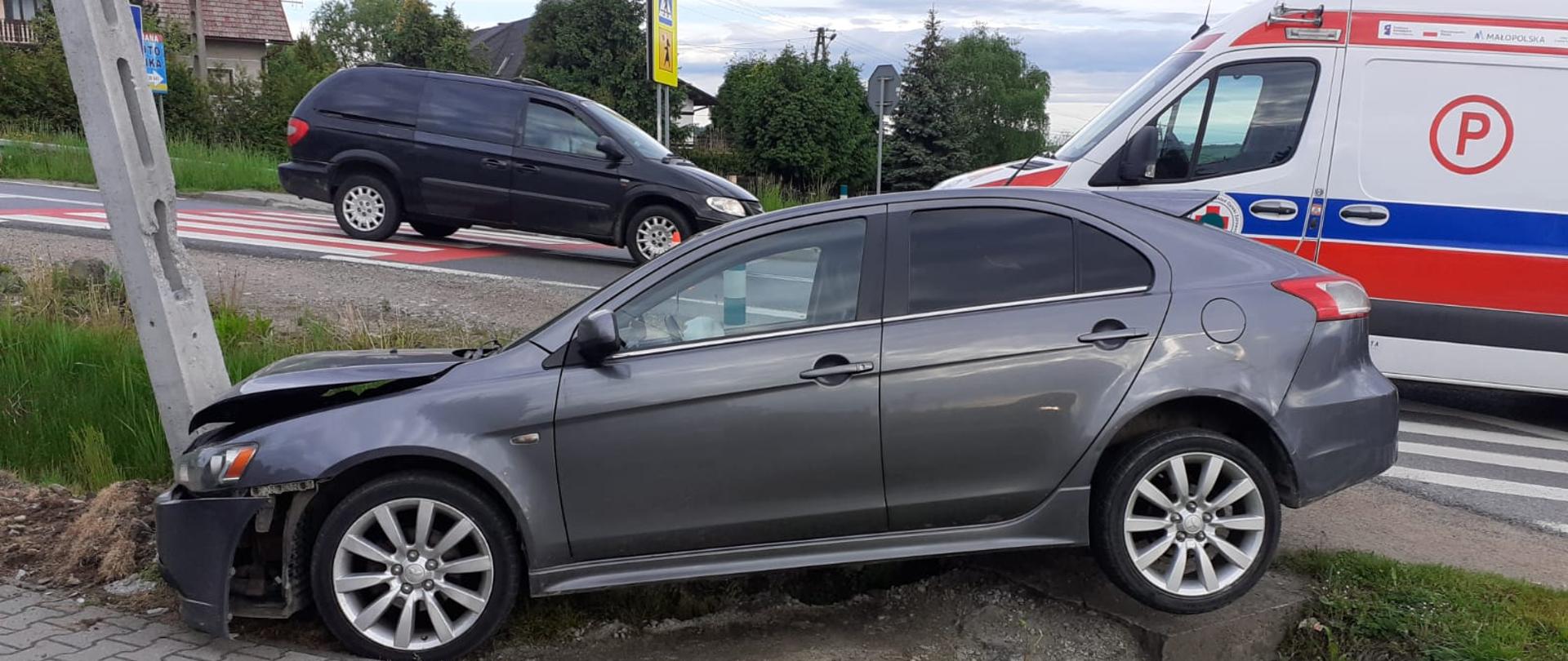 Na pierwszym planie widoczny jest samochód osobowy, który uderzył w betonowy słup. Z prawej strony na drodze stoi karetka pogotowia. Z lewej strony widzimy samochód omijający miejsce zdarzenia. W tle znajdują się zabudowania miejscowości. 