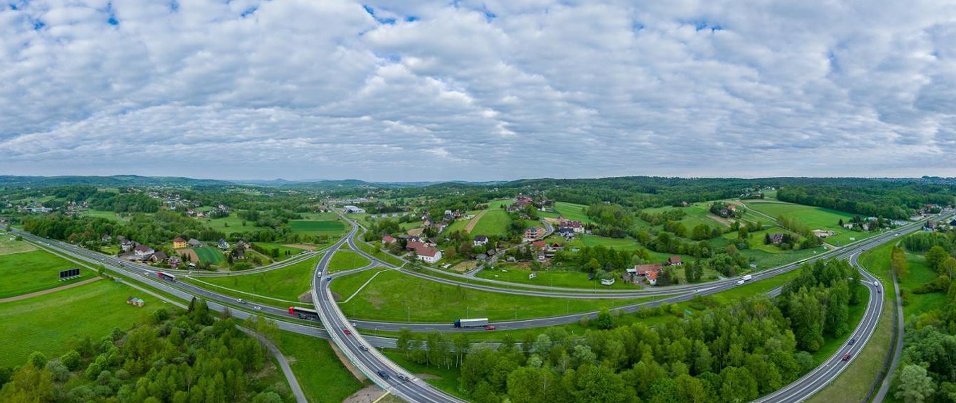 Panoramiczne zdjęcie lotnicze rozbudowanego węzła drogowego na terenach podmiejskich. Kilkupasmowe drogi i estakada przecinają zielone pola, zadrzewienia oraz rozproszoną zabudowę jednorodzinną. Na jezdniach widoczne są samochody osobowe i ciężarowe. W tle rozciągają się łagodne wzgórza i lasy, a nad krajobrazem znajduje się rozległe, częściowo zachmurzone niebo.