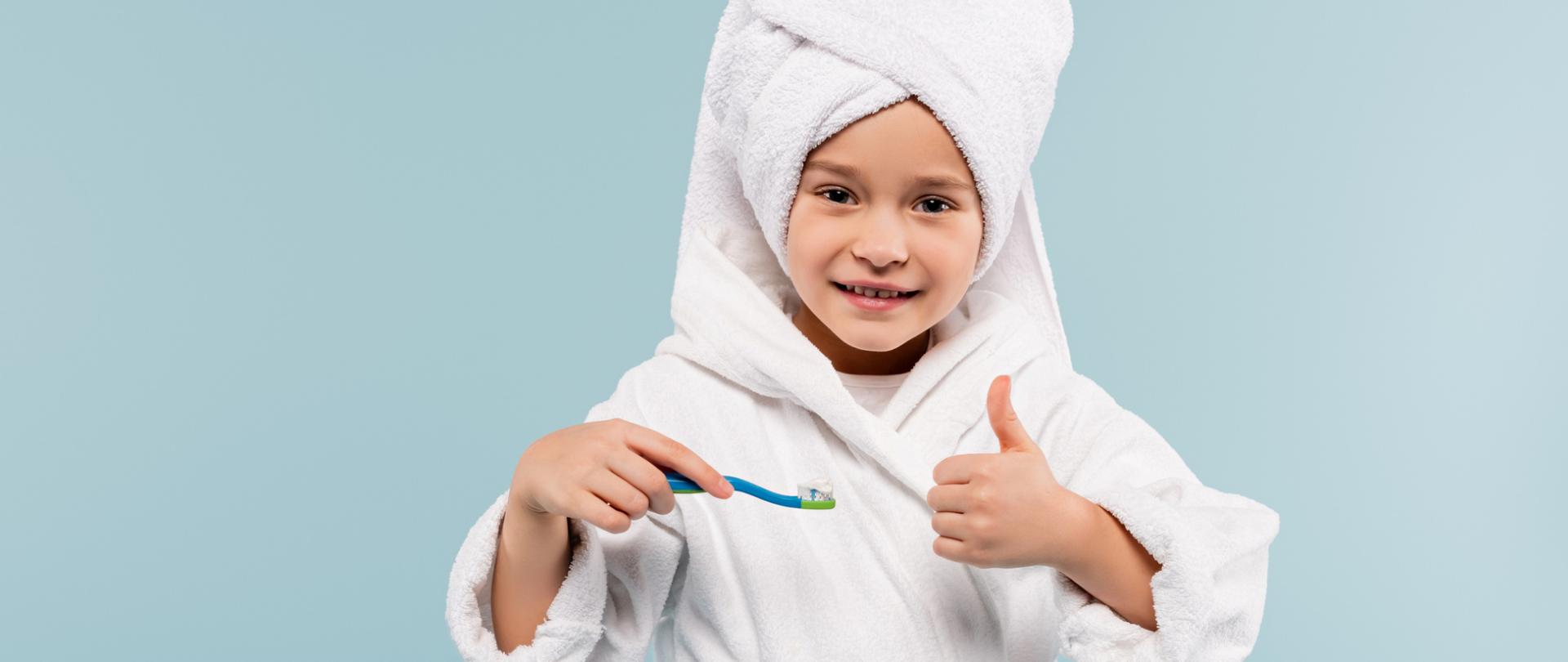 Na zdjęciu jest chłopczyk, który w reku trzyma szczoteczkę do zębów. Tło jest niebieskie.