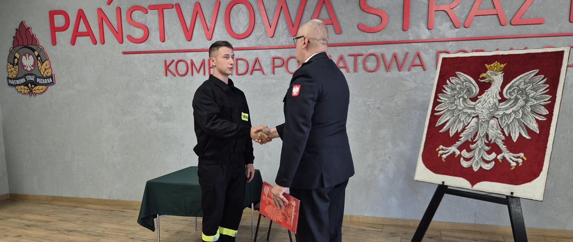 Komendanta, który składa gratulację ślubowanemu strażakowi. Obydwoje ściskają swoją prawe dłonie wzajemnie.