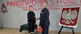 Komendanta, który składa gratulację ślubowanemu strażakowi. Obydwoje ściskają swoją prawe dłonie wzajemnie.
