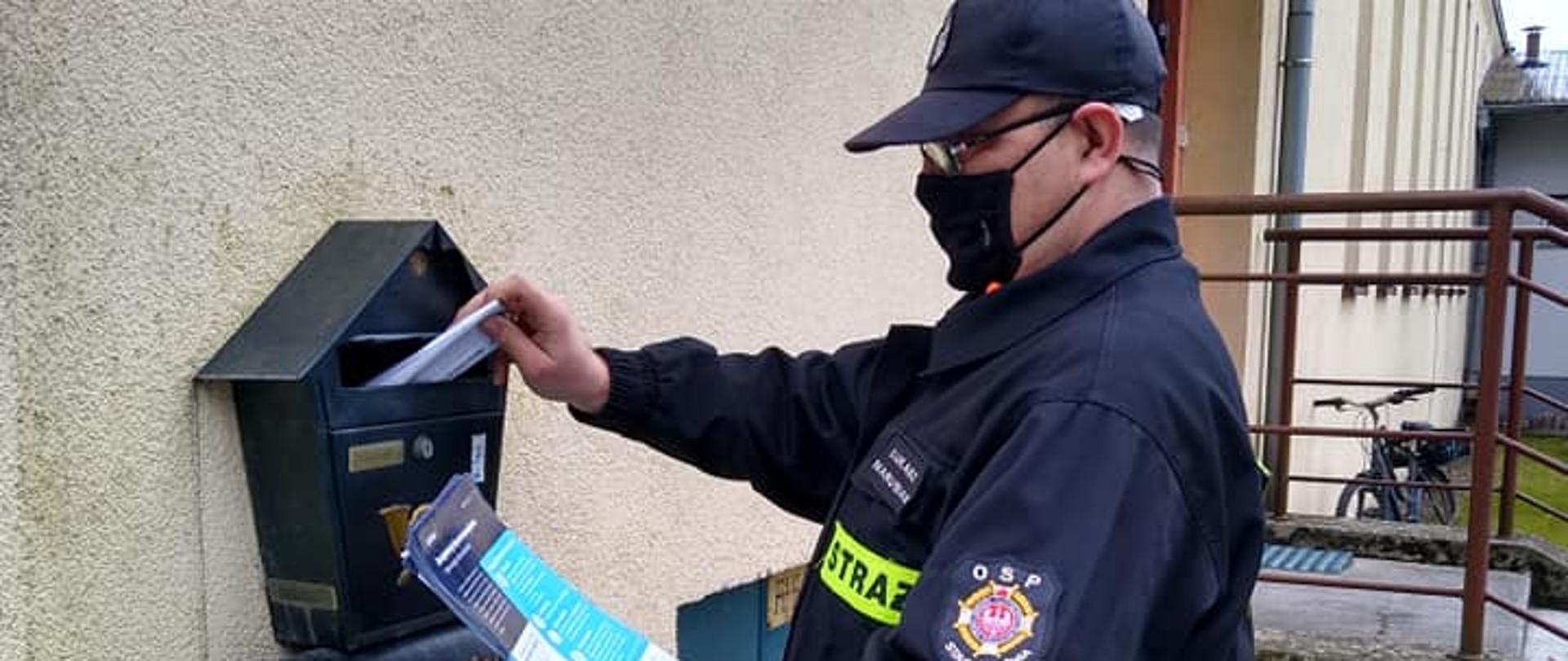 Strażak OSP w czapce i maseczce wrzucva ulotkę do skrzynki pocztowej