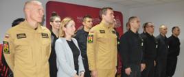 Wręczenie listu gratulacyjnego. Zdjęcie przedstawia świetlicę komendy. Na tle ścianki promocyjnej w dwuszeregu stoja strażacy i pracownica cywilna komendy. Uczestniczą w okolicznościowej zbiórce. Dwóch ze strażaków ma na sobie mundury w kolorze piaskowym, pozostali - czarne. Trzy kobiety wystepuja w strojach cywilnych.