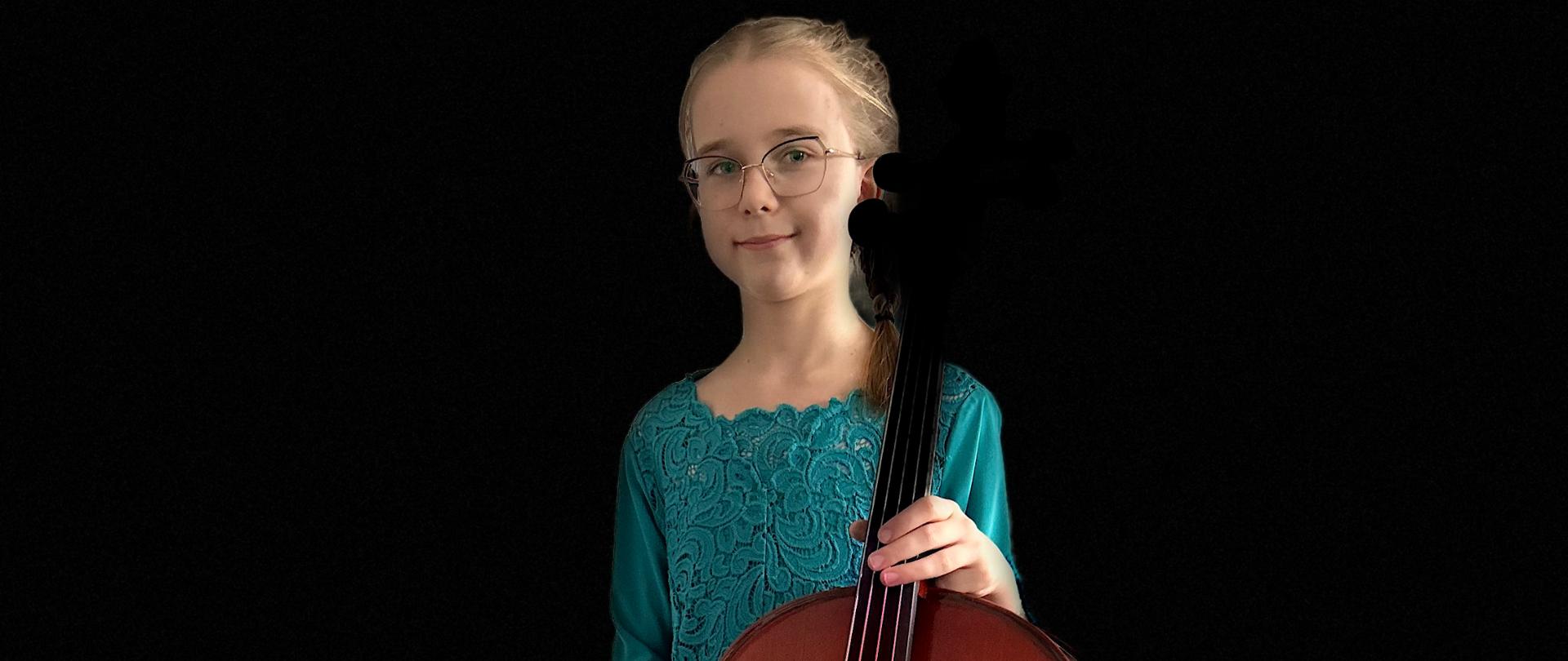 Uśmiechnięta dziewczynka w okularach ubrana w turkusową sukienkę trzyma wiolonczelę. 