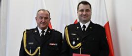 Dwóch strażaków w mundurach wyjściowych ze sznurem stoi obok siebie jeden z nich trzyma czerwoną teczkę z orłem za nimi ustawione są trzy flagi Polski.