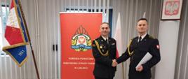 Zdjęcie zrobione w pomieszczeniu strażnicy na tle na tle Sztandaru Komendy Powiatowej Państwowej Straży Pożarnej w Lublińcu, dwóch Flag Rzeczpospolitej Polskiej oraz baneru Komendy Powiatowej PSP w Lublińcu. Na pierwszym planie Komendant Powiatowy Państwowej Straży Pożarnej w Lublińcu st. bryg. Janusz Bula z uściskiem dłoni z funkcjonariuszem odchodzącym na zaopatrzenie emerytalne. Strażacy w umundurowaniu galowym.