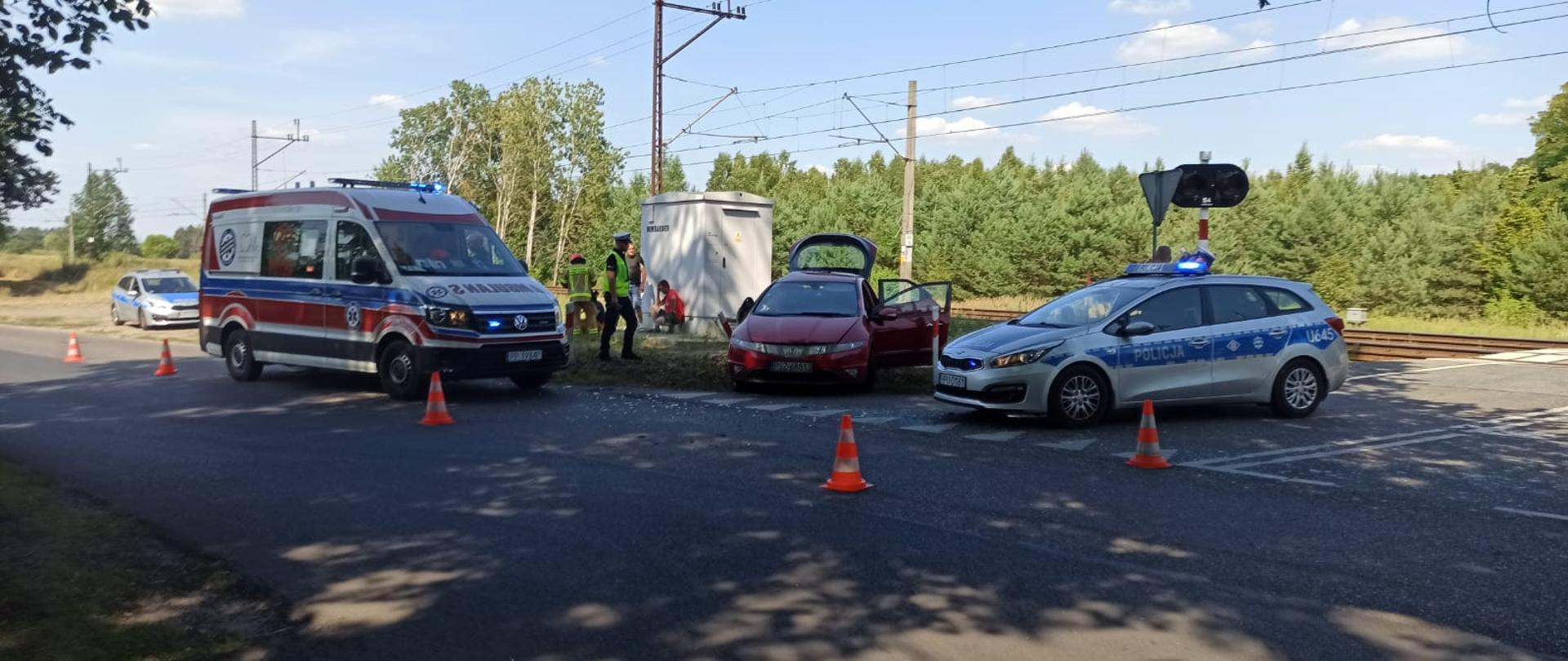 Dzień, słonecznie, bez opadów. Na zdjęciu widnieje samochód po wypadku w kolorze czerwonym oraz radiowóz Policji i karetka pogotowia ratunkowego. 