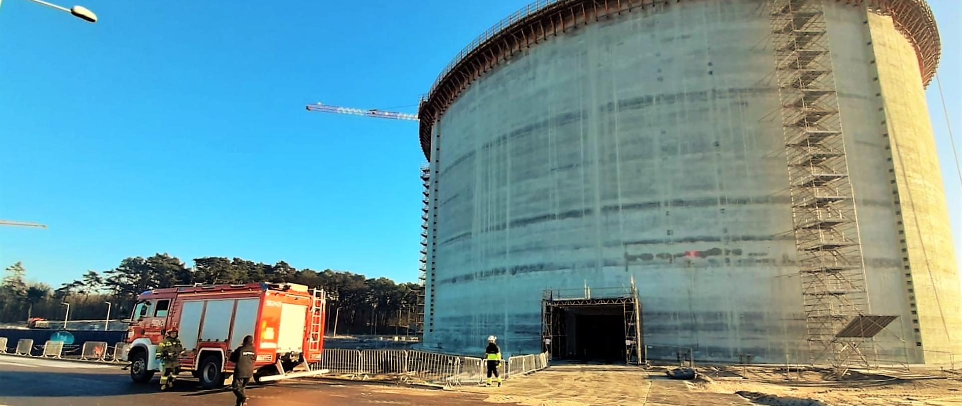 Teren terminalu LNG. Zbiornik gazu w trakcie budowy. Przed zbiornikiem średni samochód ratowniczo – gaśniczy. Dowódca zastępu strażaków udaje się w kierunku pracownika terminalu w celu ustalenia szczegółów zdarzenia.