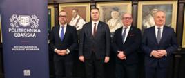 Minister Czarnek i trzech mężczyzn w garniturach stoją na tle ściany, na której wiszą portrety w złoconych ramach. Obok stoi baner z napisem Politechnika Gdańska.