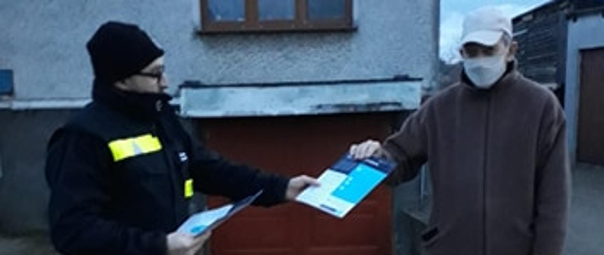 NA ZDJĘCIU WIDAC 2 MĘŻCZYZN. JEDEN Z NICH TO STRAŻAK W DŁONIACH TRZYMA PAPIEROWE ULOTKI I PRZEKAZUJE JE DRUGIEMU MĘŻCZYŹNIE.