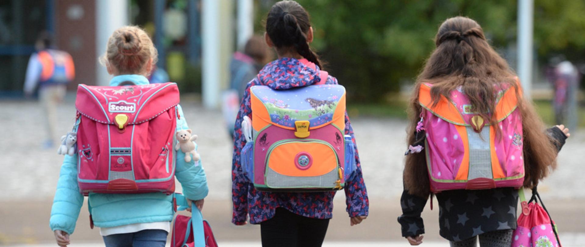 Na zdjęciu widoczne trzy dziewczynki z plecakami na ramionach. Dziewczynki ubrane w kurki (niebieską, we wzorki fioletowo różowe i szarą w gwiazdki koloru siwego). Dziewczyna z lewej - blondyna z upiętymi włosami, po środku brunetka uczesana w warkocz, który lezy na prawym ramieniu. Ostatnia dziewczynka szatynka ma włosy. długie, rozpuszczone, które leżą na plecaku.