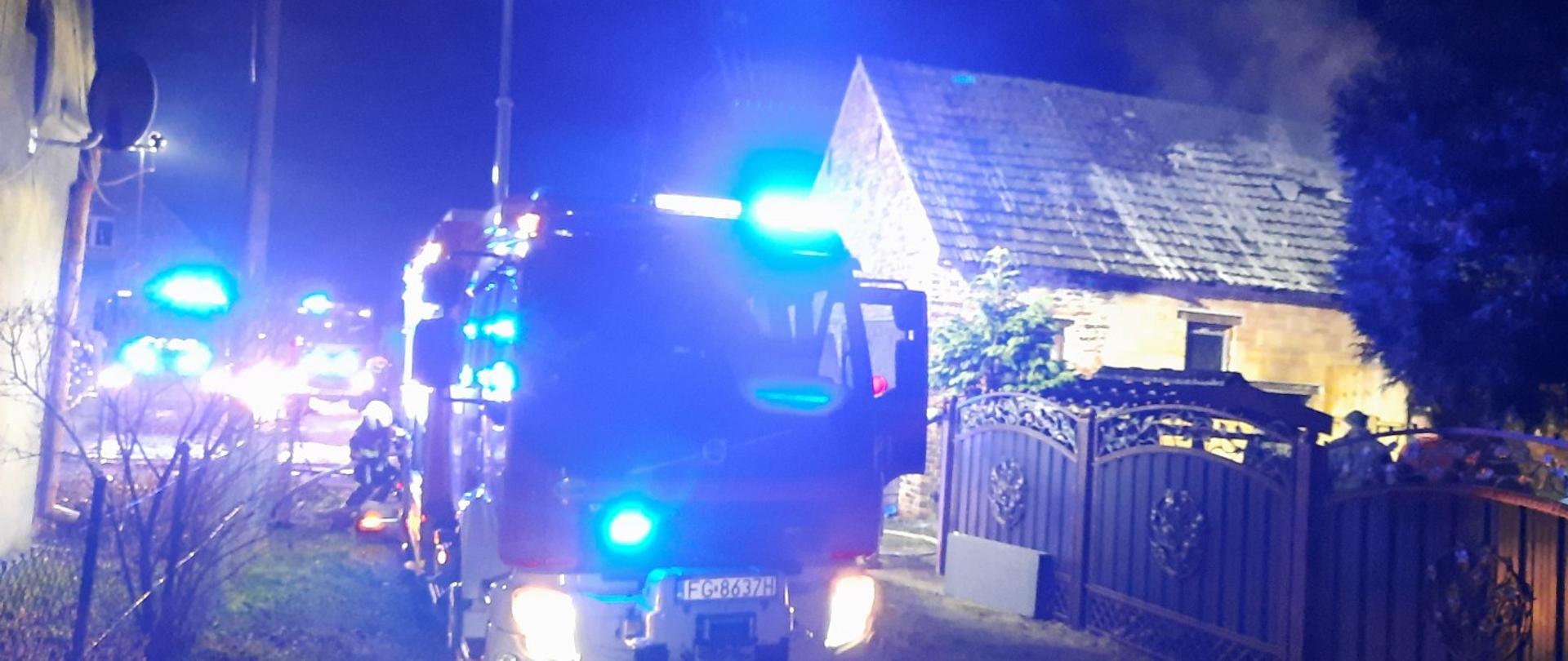 Trwa akcja gaśnicza. Samochód strażacki stoi przed posesją na której powstał pożar stodoły. Z samochodu rozwinięte są linie gaśnicze, węże pożarnicze.