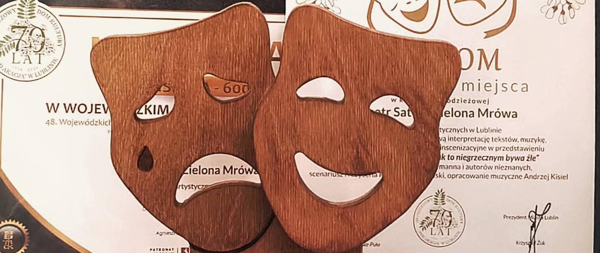 Na tle dwóch białych dyplomów z brązowym tekstem stoi drewniana statuetka. Przedstawia symbolicznie dwie maski teatralne. Na tabliczce pod spodem jest napis "Wojewódzki Przegląd Teatrów NAGRODA W KATEGORII MŁODZIEŻOWEJ Młodzieżowy Dom Kultury "Pod Akacją" Lublin 2026