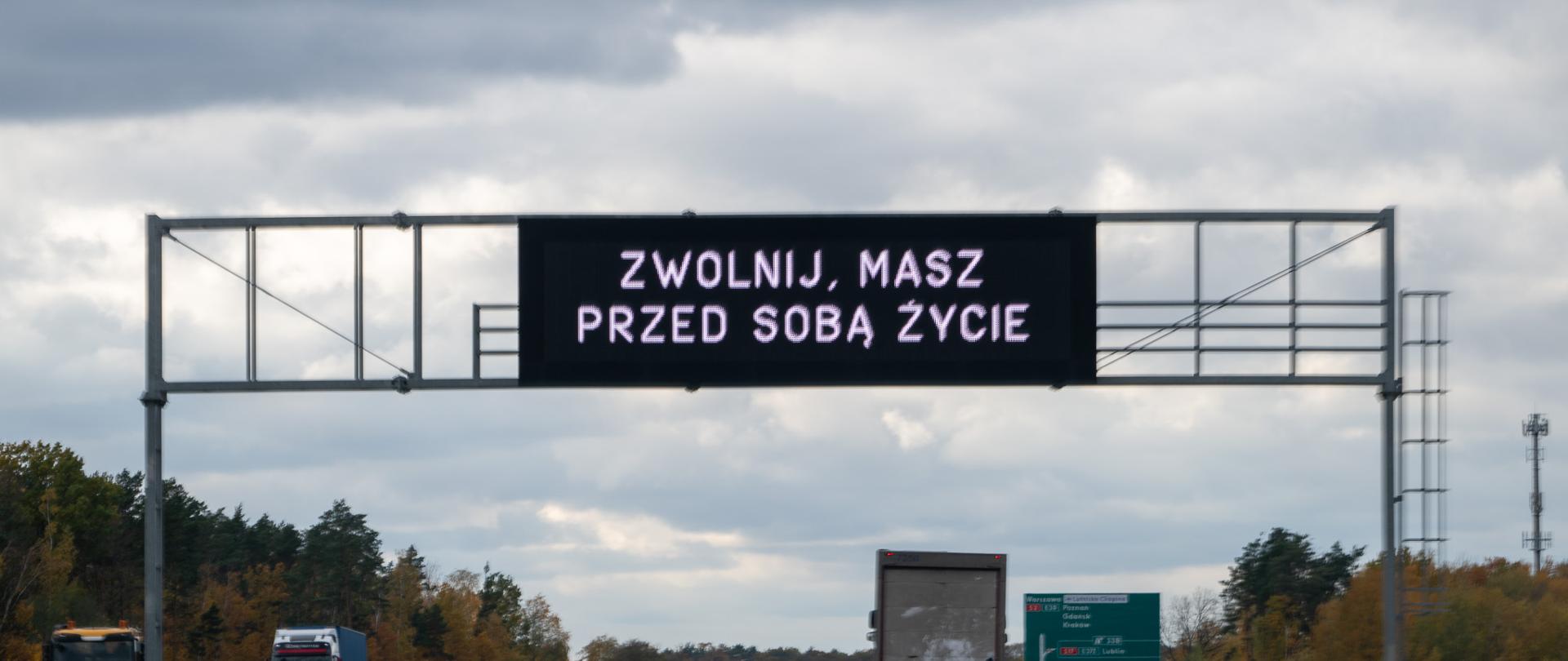 Na zdjęciu widoczny jest odcinek drogi szybkiego ruchu z kilkoma pojazdami poruszającymi się w obu kierunkach. Nad jezdnią znajduje się elektroniczna tablica informacyjna z komunikatem w języku polskim: „ZWOLNIJ, MASZ PRZED SOBĄ ŻYCIE”, który ma charakter ostrzegawczo-prewencyjny i zachęca kierowców do bezpiecznej jazdy. Po obu stronach drogi znajdują się barierki ochronne, a w tle widoczne są drzewa w jesiennych kolorach oraz tablice drogowe z informacjami kierunkowymi. Niebo jest częściowo zachmurzone, co sugeruje umiarkowane warunki pogodowe. 