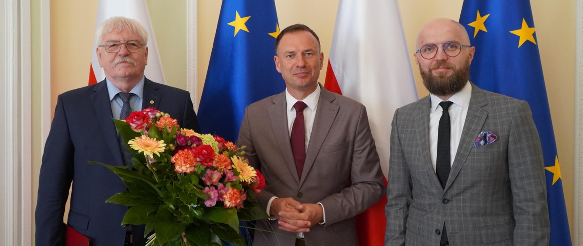 Marek Drob nowy dyrektorem Instytutu Sportu. Stoi po prawej stronie w garniturze oraz krawacie na tle flagi Unii Europejskiej. Na zdjęciu po lewej stronie stoi wiceminister Piotr Borys oraz były dyrektor Jerzy Eliasz z bukietem kwiatów w dłoni
