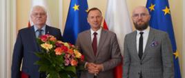 Marek Drob nowy dyrektorem Instytutu Sportu. Stoi po prawej stronie w garniturze oraz krawacie na tle flagi Unii Europejskiej. Na zdjęciu po lewej stronie stoi wiceminister Piotr Borys oraz były dyrektor Jerzy Eliasz z bukietem kwiatów w dłoni
