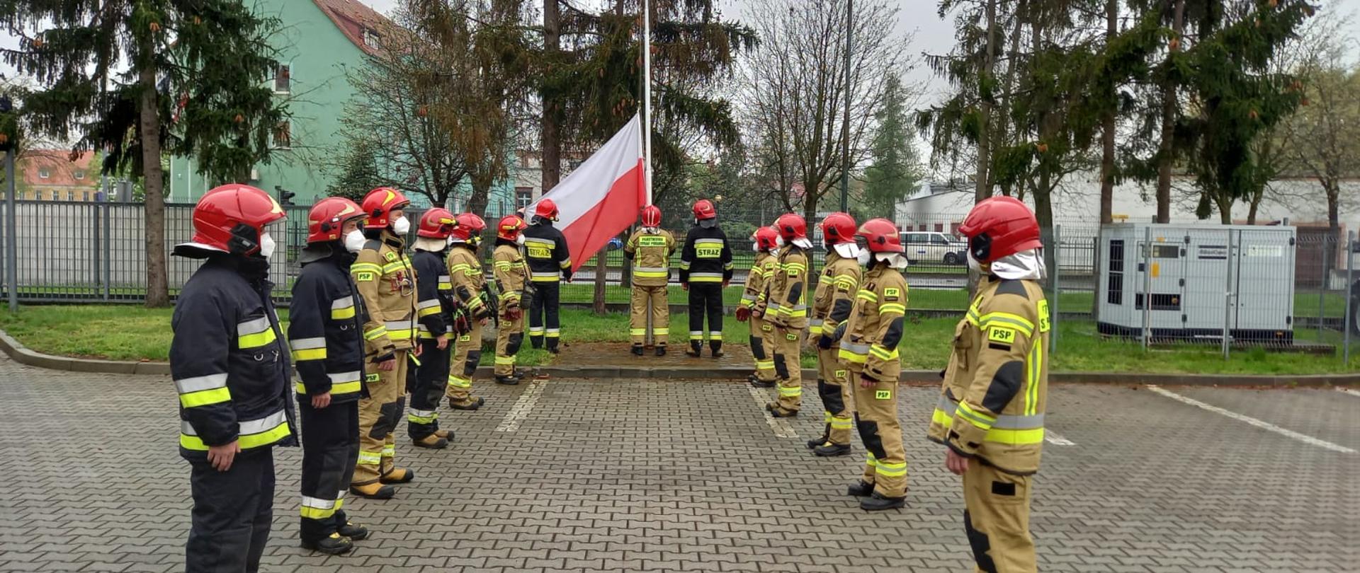 Strażacy w umundurowaniu specjalnym stojący w dwóch szeregach na przeciwko siebie. W tle maszt flagowy z zawieszoną u dołu flagą Polski i stojący przed masztem poczet flagowy.