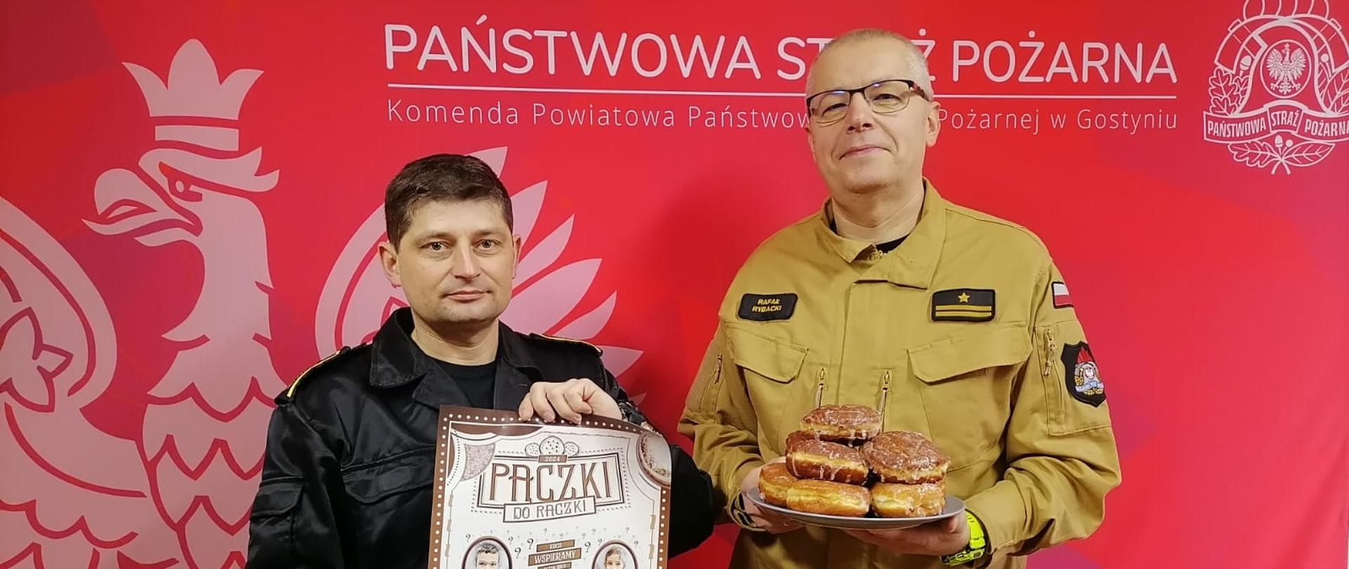 Dwóch strażaków stojących i pozujących do zdjęcia. Jeden z nich trzyma w ręce plakat akcji pączki do rączki. Drugi trzyma w ręce talerz z pączkami.